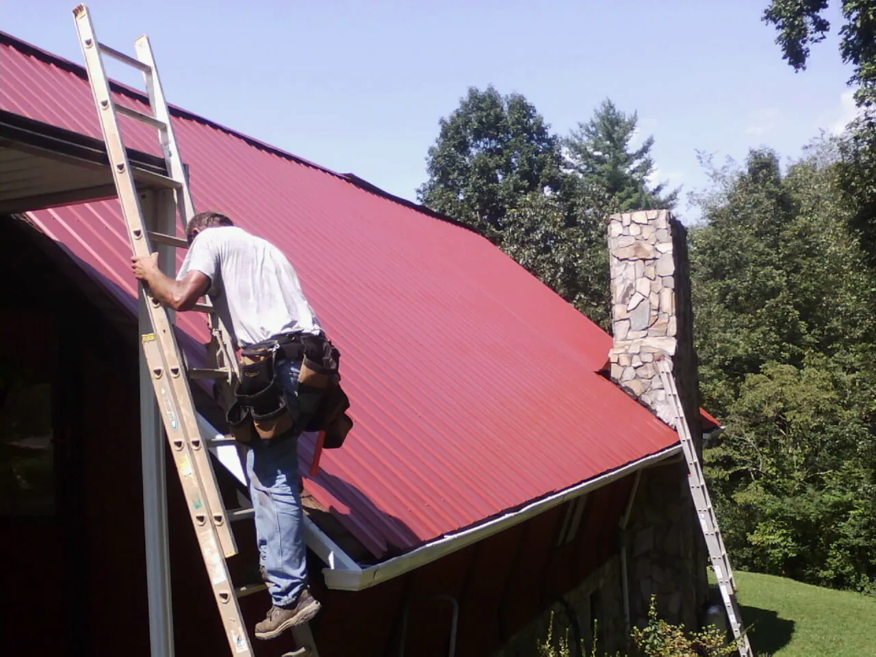 Roofer on a ladder beside a red metal roof and stone chimney — Soffit & Fascia Repair by Roofing Brothers in Mercersburg